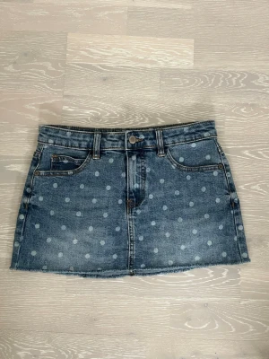 Blå prickig jeanskjol  - Snygg blå jeanskjol med ljusa prickar och rå kant nertill. Kjolen har klassisk femficksmodell, bälteshällor och knappstängning fram. Perfekt för dig som gillar denim med en twist.