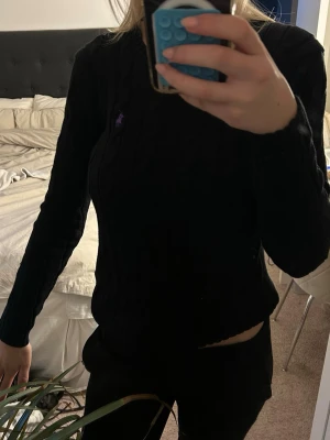 Svart kabelstickad tröja Polo Ralph Lauren - Kabelstickad svart tröja från Polo Ralph Lauren i mjuk pima cotton. Klassisk rund halsringning och lång ärm. Lila broderad logga på bröstet ger en snygg kontrast. Perfekt till jeans eller kjol för en clean look.