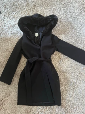 Svart kappa med fuskpäls från H&M - Stilren svart kappa från H&M med stor huva kantad av mjuk fuskpäls. Kappan har knytband i midjan och raka ärmar. Perfekt för kalla dagar när du vill vara både varm och snygg. Klassisk design som passar till många olika outfits.