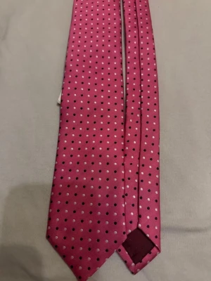 Rosa slips med prickigt mönster - En snygg rosa slips med små svarta prickar över hela ytan. Slipsen har klassisk form och är tillverkad i ett glansigt material som känns lyxigt. Perfekt för dig som vill sticka ut med en färgglad accessoar.