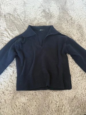 Mörkblå stickad tröja från Gina Tricot - Snygg mörkblå stickad tröja från Gina Tricot med krage och v-ringning. Tröjan har långa ärmar och bred ribbstickad kant nedtill samt vid ärmsluten. Perfekt för en stilren och enkel look.