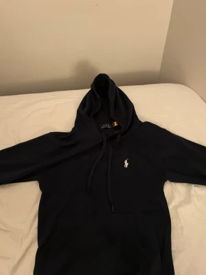 Mörkblå hoodie  från Polo Ralph Lauren - Klassisk mörkblå hoodie från Polo Ralph Lauren med vit broderad logga på bröstet. Hoodien har justerbar dragsko i huvan och är tillverkad i mjukt material som känns skönt mot huden. Perfekt för en avslappnad och stilren look.