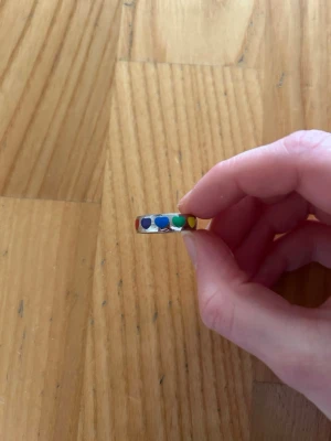 Färgglad ring med hjärtan - Säljer en silverfärgad ring dekorerad med små hjärtan i olika färger runt hela ringen. Hjärtana är i rött, blått, grönt, gult och orange. Ringen har en slät och enkel form som gör den lätt att matcha med andra smycken.