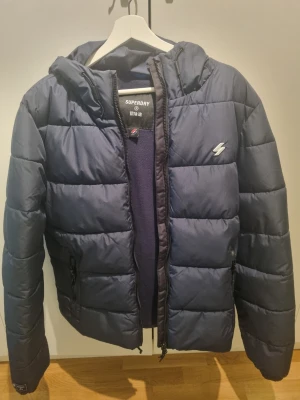 Mörkblå pufferjacka från Superdry - Snygg mörkblå pufferjacka från Superdry med huva och dragkedja framtill. Jackan har två sidofickor med dragkedja och ett diskret Superdry-märke på bröstet. Perfekt för kalla dagar tack vare den vadderade designen och den sportiga looken.
