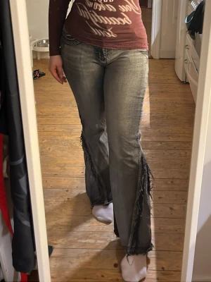 Grå slitna bootcut jeans med fransar - Säljer ett par grå bootcut jeans med slitna detaljer och fransar längs sidorna. Jeansen har klassisk femficksmodell och markerade sömmar. Materialet är denim med tvättad look och de har en avslappnad passform som ger en cool vibe.