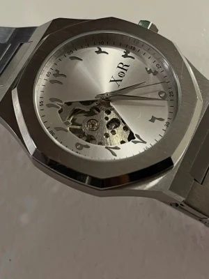 XoR Obsucra 41mm Silver/Grå HELT NY - Stilren klocka från XoR i borstat silverfärgat 316L rostfritt stål med unikt urverk synligt på urtavlan. Klockan har arabiska siffror, rund urtavla och länkarmband i metall. Kommer helt ny med originalbox. Plasten är på eftersom den är sprillans ny.