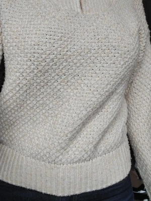 Beige stickad tröja med halv dragkedja - Mysig beige stickad tröja med halv dragkedja vid halsen och ribbade muddar. Tröjan har ett diskret strukturmönster och är perfekt för kyliga dagar. Passar till jeans eller kjol för en avslappnad look.