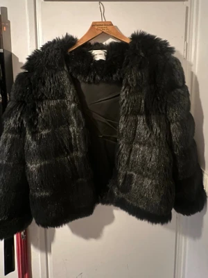 Svart fuskpälsjacka från Dry Lake - Snygg svart fuskpälsjacka från Dry Lake med fluffig krage och breda ärmar. Jackan har en glansig finish och är helfodrad för extra värme. Perfekt för att hålla stilen under kalla dagar. De står att jackan är i s men passar även XS och S