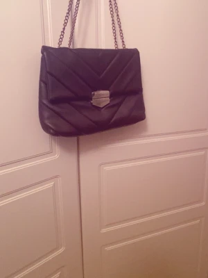 Svart quiltad axelväska med kedja - Snygg svart axelväska i quiltat mönster med kedjerem och silverfärgat spänne framtill. Väskan har en klassisk rektangulär form.