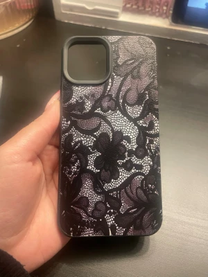 Svart mobilskal med spetsmönster - Snyggt mobilskal till iPhone 12 Pro med svart och vitt spetsinspirerat blommönster. Skalet är gjort i tålig plast och har en matt finish som ger ett elegant intryck. Perfekt för dig som vill ha ett unikt och stilrent skydd till mobilen.