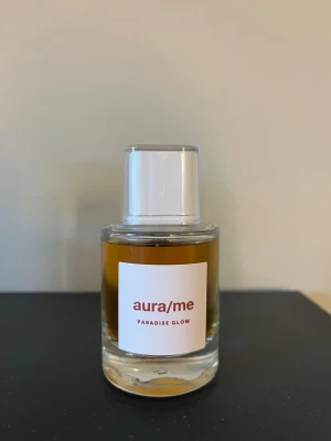 aura/me Paradise Glow parfym - Parfymflaska från aura/me med doften Paradise Glow. Flaskan är cylindrisk i klart glas med vit etikett och vit plastkork. Innehållet är bärnstensfärgat vilket ger en lyxig känsla. Perfekt för dig som vill ha en unik och modern doft i samlingen.