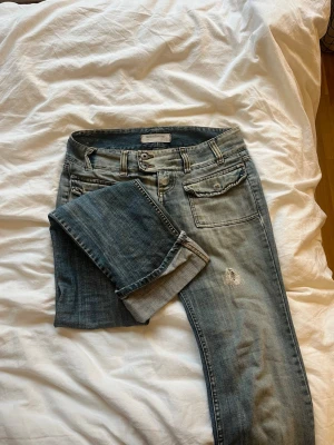 Low waist bootcut jeans med hål i  - Säljer ett par blå jeans med snyggt slitna detaljer och dubbla knappar i midjan. Jeansen har fem fickor, raka ben och slitningar både fram och bak, säljer pga försmå😄