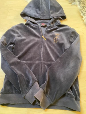 Grå velourhoodie från Juicy Couture - Mjuk och lyxig grå velourhoodie från Juicy Couture med dragkedja framtill. Har huva, ribbade muddar och guldfärgade strasstenar som bildar logga på bröstet och stor Juicy Couture-text på ryggen. Perfekt för en avslappnad och trendig look.