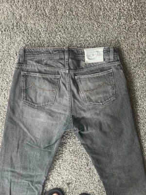 Gråa jeans från Jacob Cohen - Säljer ett par gråa jeans från Jacob cohen med raka ben och klassiska bakfickor. Jeansen har en diskret tvättad look och kontrastsömmar på fickorna. Materialet är denim i bomull och passformen är normal. Perfekta för en clean och stilren outfit. Storleken är 32 men sitter mer som 30