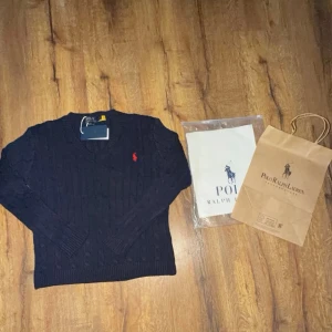 Mörkblå kabelstickad tröja från Polo Ralph Lauren - Snygg mörkblå kabelstickad tröja från Polo Ralph Lauren med röd broderad logga på bröstet. Tröjan har V-ringning, långa ärmar och ribbade muddar. Perfekt för dig som gillar klassisk och stilren design. Med tags för bara 800 kr priset går att diskuteras vid snabb affär ny pris 2500 kr