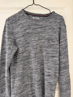 Gråmelerad tröja från Tommy Hilfiger - Snygg gråmelerad långärmad tröja från Tommy Hilfiger med diskret logga på bröstet. Tröjan har rund halsringning och är tillverkad i mjukt material som passar perfekt till en avslappnad stil.