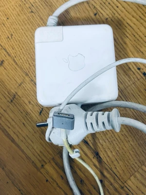 Apple MagSafe laddare till MacBook - Vit MagSafe-laddare från Apple för MacBook. Fyrkantig form med Apple-logga på ovansidan och lång kabel med MagSafe-kontakt. Tillverkad i plast och gummi, passar till äldre MacBook-modeller.