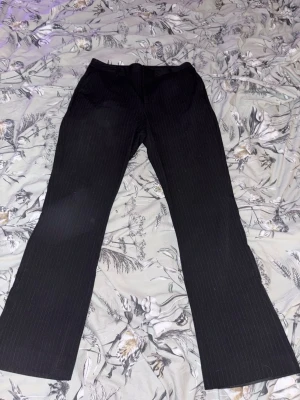 Svarta kostymbyxor med smala ränder - Snygga svarta highwaisted kostymbyxor med diskreta vita kritstrecksränder. Byxorna har raka ben och klassisk passform, perfekta för en stilren look. Materialet känns mjukt och följsamt, troligen en blandning av polyester och viskos. 