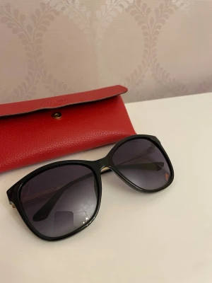 Svarta Guess solglasögon med fodral - Snygga solglasögon från Guess med svart båge och guldfärgade detaljer på skalmarna. Dock lite skav på se bilder.  Kommer med ett rött fodral i läderlook. Perfekta för att höja din stil i solen.