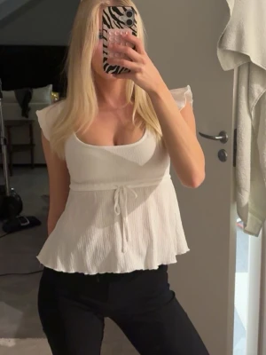 Vit ribbad linnetopp med knyt - Söt vit linnetopp med ribbad struktur och volangkant. Toppen har korta ärmar, rundad urringning och knyt i midjan för en feminin siluett. Perfekt till jeans eller kjol för en fräsch look.