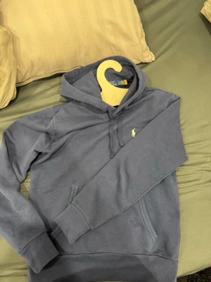 Blå hoodie Polo Ralph Lauren - Snygg blå hoodie från Polo Ralph Lauren med klassisk logga broderad på bröstet. Hoodien har huva med dragsko och en stor magficka framtill. Tillverkad i mjukt material som känns skönt mot huden. Perfekt för en avslappnad och stilren look.