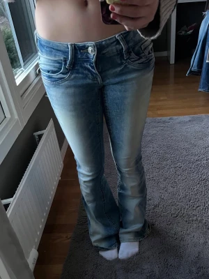 Blå bootcut jeans med slitningar - Säljer ett par ljusblå bootcut jeans med snygga slitningar och tvättade detaljer. Jeansen har låg midja, klassiska fem fickor och stängs med knapp och dragkedja. Perfekta för en avslappnad och trendig look.