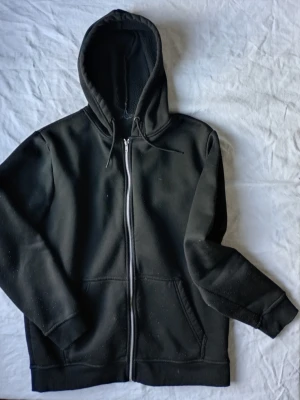 Svart zip hoodie med huva - Klassisk svart hoodie med dragkedja framtill och stor huva. Hoodien har två fickor på magen och ribbade muddar vid ärmslut och nederkant. Perfekt för en avslappnad och stilren look. Materialet känns mjukt och bekvämt.