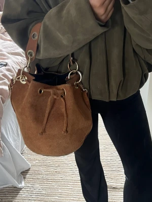 Brun mockaväska med snörning - Säljer en snygg brun bucket bag i mocka med snörning och guldfärgade metalldetaljer. Väskan har både ett kortare handtag och avtagbart axelband som man kan anpassa i längd. Väskan är handgjord😍 villig att förhandla priset