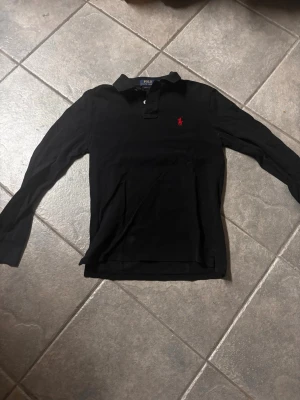Svart långärmad polotopp från Polo Ralph Lauren - Klassisk svart långärmad polotopp från Polo Ralph Lauren med krage och knappar framtill. Ikoniska röda loggan broderad på bröstet. Tillverkad i mjuk bomull som känns skön mot huden. Perfekt till jeans eller kjol för en clean och stilren look.