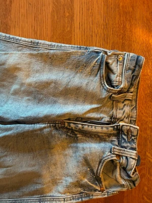 Ljusblå jeans  - Säljer ett par ljusblå jeans med dekorativt glitter vid fickorna. Jeansen har klassisk femficksmodell, dragkedjegylf och bälteshällor. 