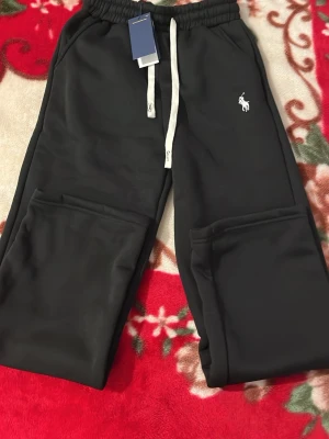 Svarta mjukisbyxor från Polo Ralph Lauren - Snygga svarta mjukisbyxor från Polo Ralph Lauren med vit dragsko i midjan och klassisk logga broderad på vänster sida. Byxorna har resår i midjan och muddar vid bensluten. Perfekta för chill dagar och streetwear-stil.