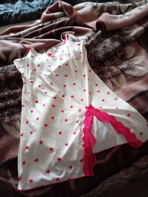 Vitt nattlinne med rosa hjärtan - Sött vitt nattlinne med tunna axelband och rosa hjärtmönster. Nattlinnet har en dekorativ rosa spetskant längs nederdelen och en liten slits framtill. Perfekt för mysiga kvällar hemma.stl L/XL
