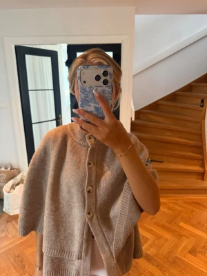 Beige stickad cape med knappar - Säljer en beige stickad cape med stora träknappar framtill. Capen har en lös passform, ribbade kanter och hög krage. Perfekt för lager på lager och ger en chill men ändå stilren look.