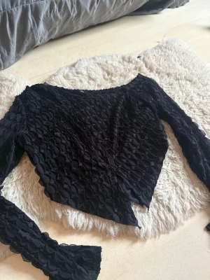 Svart asymmetrisk topp från SHEIN - Trendig svart topp från SHEIN med lång ärm och asymmetrisk nederkant. Toppen har ett unikt strukturerat mönster och är tillverkad i ett syntetmaterial. Perfekt för dig som vill sticka ut med en cool och modern look.