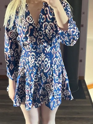 Blå mönstrad kortklänning - Säljer en blå kortklänning med vitt och rosa mönster. Klänningen har v-ringad hals och långa ärmar. Kjolen är volangig och ger en flowy känsla. Perfekt för sommarens alla tillfällen!