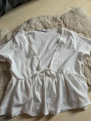 Vit omlottopp med knyt - Säljer en söt vit omlottopp med knyt framtill och korta ärmar. Toppen har en djup v-ringning och lätt rynkad midja som ger en fin siluett. Materialet är ribbat och känns mjukt mot huden. Perfekt för dig som gillar en enkel men trendig stil.