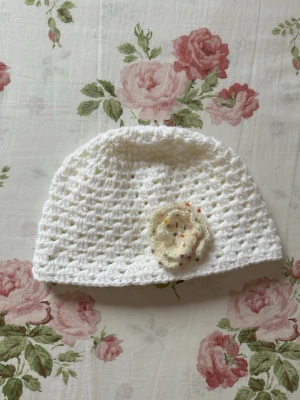 Vit virkad mössa med blomma - Söt handvirkad vit mössa med en dekorativ cremefärgad blomma framtill. Mössan har ett luftigt, hålmönstrat virkade och blomman har små färgade prickar för extra detalj. Perfekt accessoar för dig som gillar en romantisk och unik stil.
