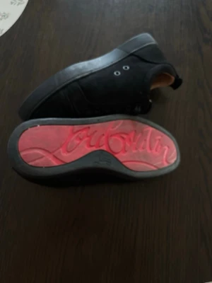 Svarta sneakers från Christian Louboutin - Snygga svarta sneakers från Christian Louboutin med klassisk röd sula och diskret logga undertill. Skorna har lågt skaft, svarta skosnören och är tillverkade i mocka. Perfekta för dig som vill ha en stilren och lyxig look.