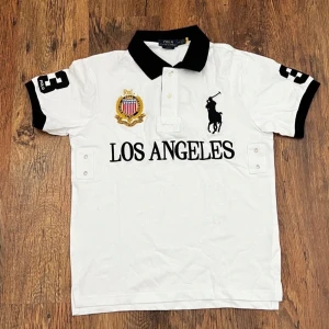 Vit polotopp från Polo Ralph Lauren - Vit polotopp med svart krage och detaljer från Polo Ralph Lauren. Stora broderade loggor på bröstet och texten 'LOS ANGELES' framtill. Svarta detaljer på ärmarna och klassisk knappslå. Perfekt för dig som gillar sportig och stilren look.
