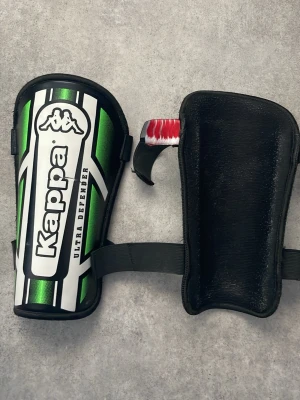 Kappa benskydd Ultra Defender  - Säljer ett par benskydd från Kappa, modell Ultra Defender. De är gröna med vita och svarta detaljer samt har justerbara kardborreband för bra passform. Perfekta för fotbollsträning eller match. Snygg och sportig design.