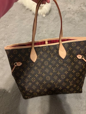 damväska - Säljer en klassisk handväska i brunt monogramcanvas med beige läderdetaljer och rödrandigt foder. Väskan har två långa handtag och öppet huvudfack, perfekt för dig som gillar lyxig och tidlös design.