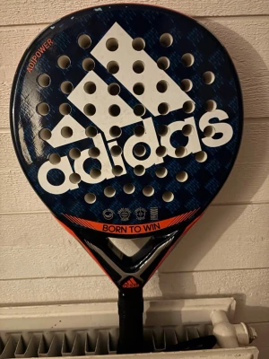 Adidas Adipower padelracket blå/orange - Säljer ett Adidas Adipower youth padelracket i blått och orange med stort vitt Adidas-tryck. Racketen har rund form, många hål och texten 'Born to win' på framsidan. Perfekt för dig som vill ta ditt padelspel till nästa nivå. 