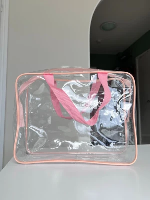 Transparent necessär med rosa detaljer - Transparent necessär i plast med rosa detaljer och dragkedja. Perfekt för smink, hudvård, hårvård, kroppsvårdsartiklar eller andra småprylar. Rektangulär form, enkel att ta med sig.