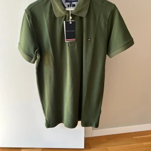 Olivgrön pikétröja Tommy Hilfiger - Snygg olivgrön pikétröja från Tommy Hilfiger med klassisk krage och korta ärmar. Tröjan har en liten broderad logga på bröstet och stängs med två knappar vid halsen. Perfekt för en clean och stilren look. Helt oanvänd 