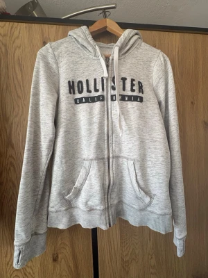 Grå zip-hoodie från Hollister - Ljusgrå hoodie med dragkedja från Hollister. Stor logga i svart text på bröstet och texten 'California' under. Utrustad med huva och vita dragsnören samt två fickor framtill. Tillverkad i mjukt sweatshirtmaterial, ribbade muddar vid ärmar och nederkant.