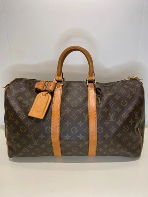 Louis Vuitton Keepall 45 Monogram - Snygg Louis Vuitton Keepall 45 resväska i klassiskt brunt monogramcanvas med detaljer i ljust läder. Väskan har två robusta handtag, guldfärgade metalldetaljer och en bagagetagg. Kommer med lås och nycklar. Perfekt storlek för weekendresor och ikonisk LV-design.