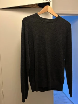 Mörkgrå stickad tröja från Uniqlo - Snygg mörkgrå stickad tröja från Uniqlo med rund halsringning och långa ärmar. Tröjan har en enkel och stilren design, perfekt att matcha med jeans eller chinos. Tillverkad i mjukt material som känns skönt mot huden. Två små hål på ryggen, kolla sista bilden 
