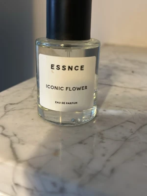 ESSNCE Iconic Flower Eau de Parfum - Fräsch parfym från ESSNCE med namnet Iconic Flower. Flaskan är cylindrisk i genomskinligt glas med svart lock och enkel vit etikett. Doften är en eau de parfum och ger en modern och stilren känsla.