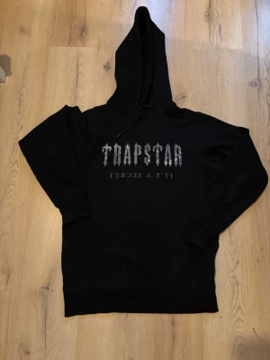 Svart Trapstar hoodie med tryck - Svart hoodie från Trapstar med stor huva och dragsko. På bröstet finns ett grått tryck med texten 'TRAPSTAR IT'S A SECRET'. Hoodien har ribbade muddar vid ärmslut och nederkant. Perfekt för en streetwear-look.