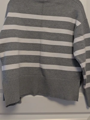 Gråvit randig stickad tröja - Säljer en stickad tröja i grått och vitt med breda horisontella ränder. Tröjan har rund halsringning, långa ärmar och ribbade muddar vid ärmslut och nederkant. 